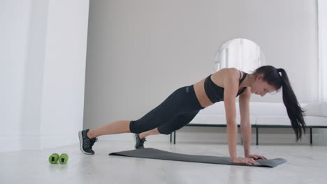 A-young-brunette-in-black-clothes-and-a-light-apartment-performs-the-plank-exercise-jumping-in-the-rest-lying-down